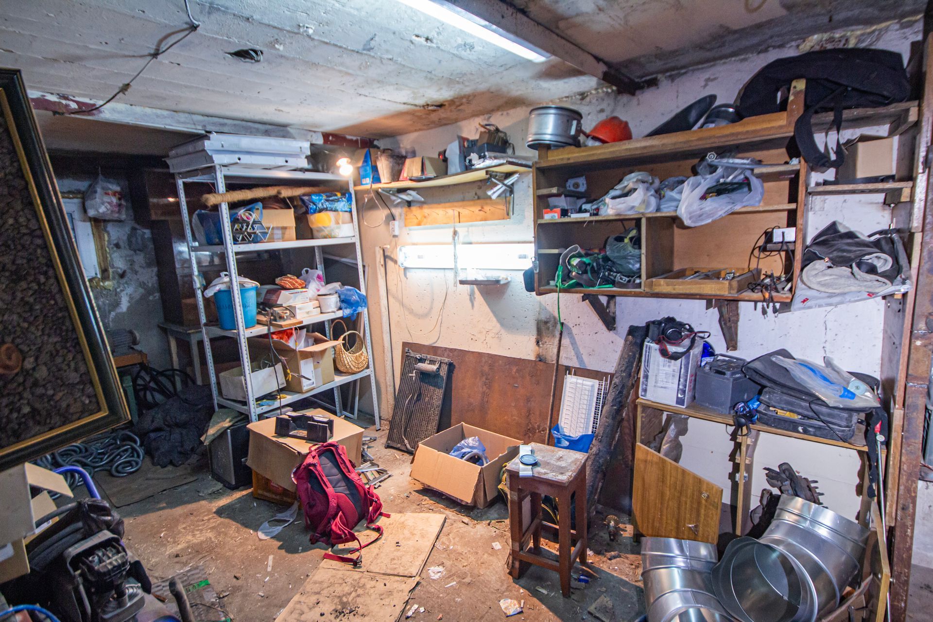 Cluttered basement storage room with shelves, boxes, and various items. Overcast lighting, dirt on floor.