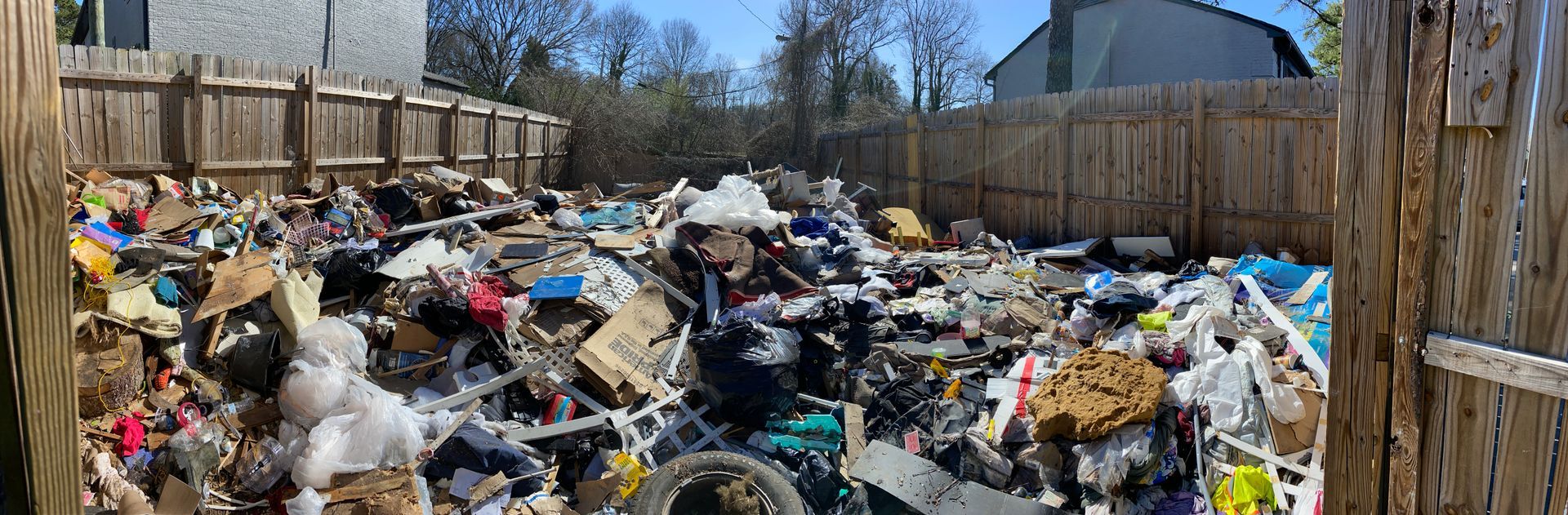 A backyard filled with a massive pile of trash and debris, surrounded by a wooden fence.