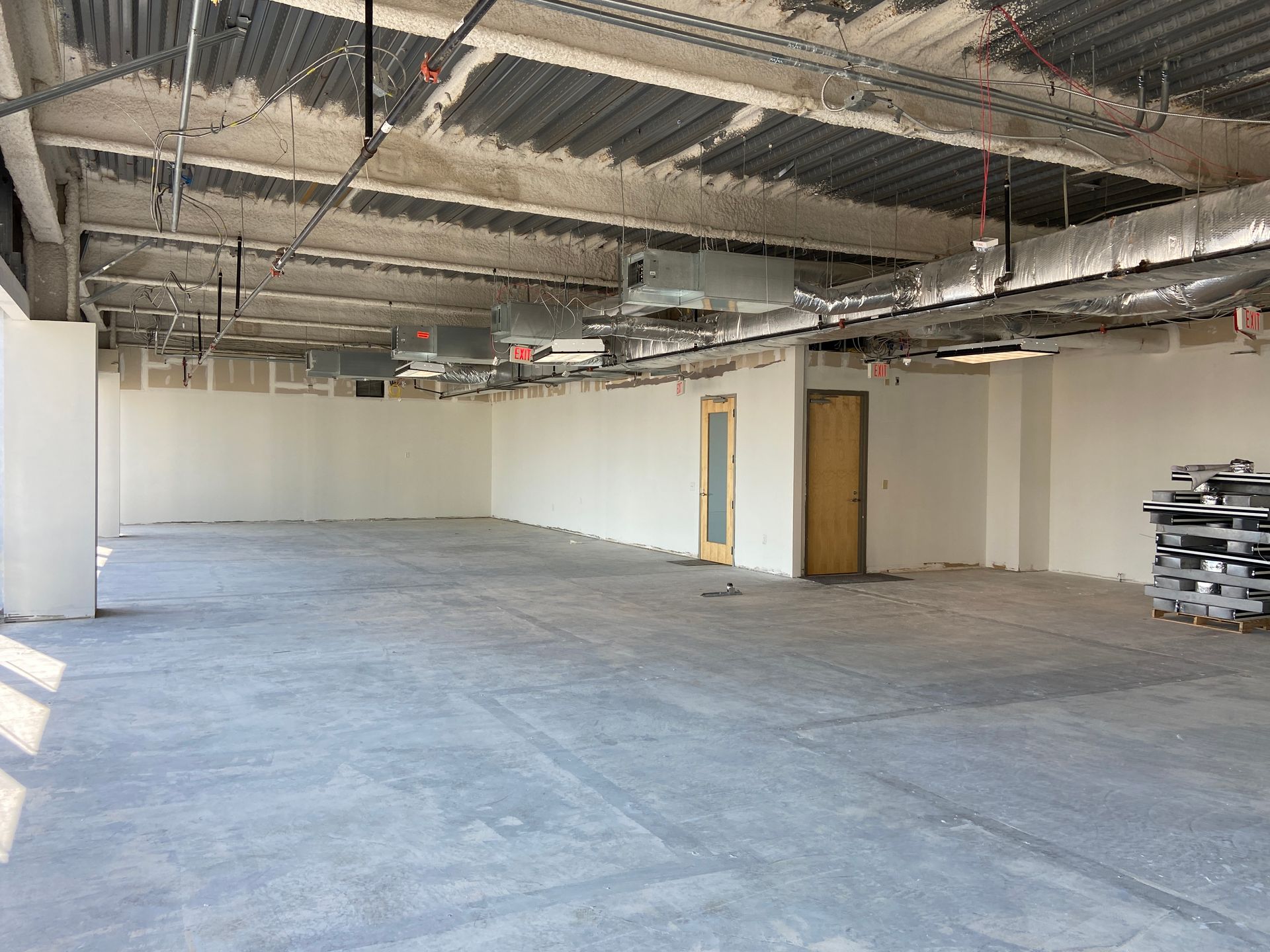 Construction site: unfinished interior room with bare concrete floor, white walls, exposed ceiling infrastructure, and a closed door.