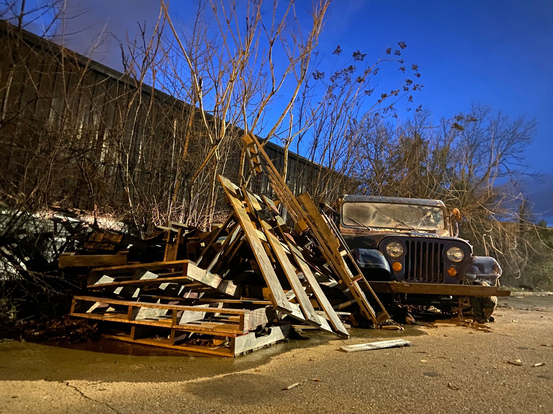 An old, rusty Jeep and wood pallets are next to a building under a blue twilight sky.