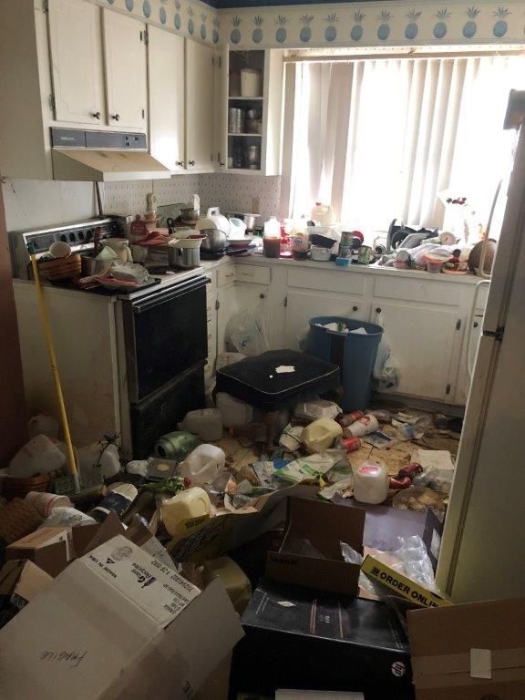 Cluttered kitchen with overflowing trash, scattered items, and dirty surfaces; the room appears unsanitary.