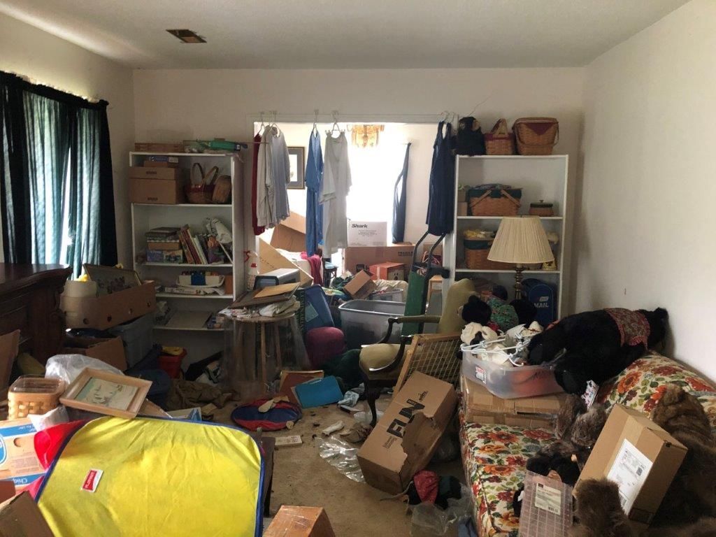 A cluttered room with boxes, furniture, and personal belongings piled up. Shelves and window are also filled.