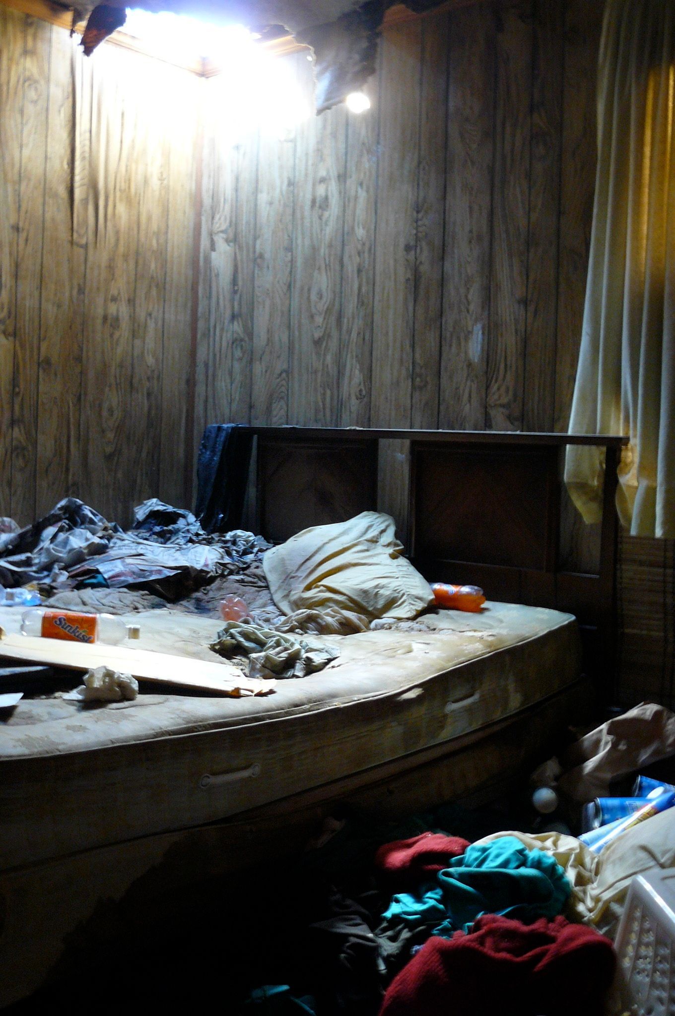 Messy room with a damaged ceiling, cluttered mattress, and piles of clothes.
