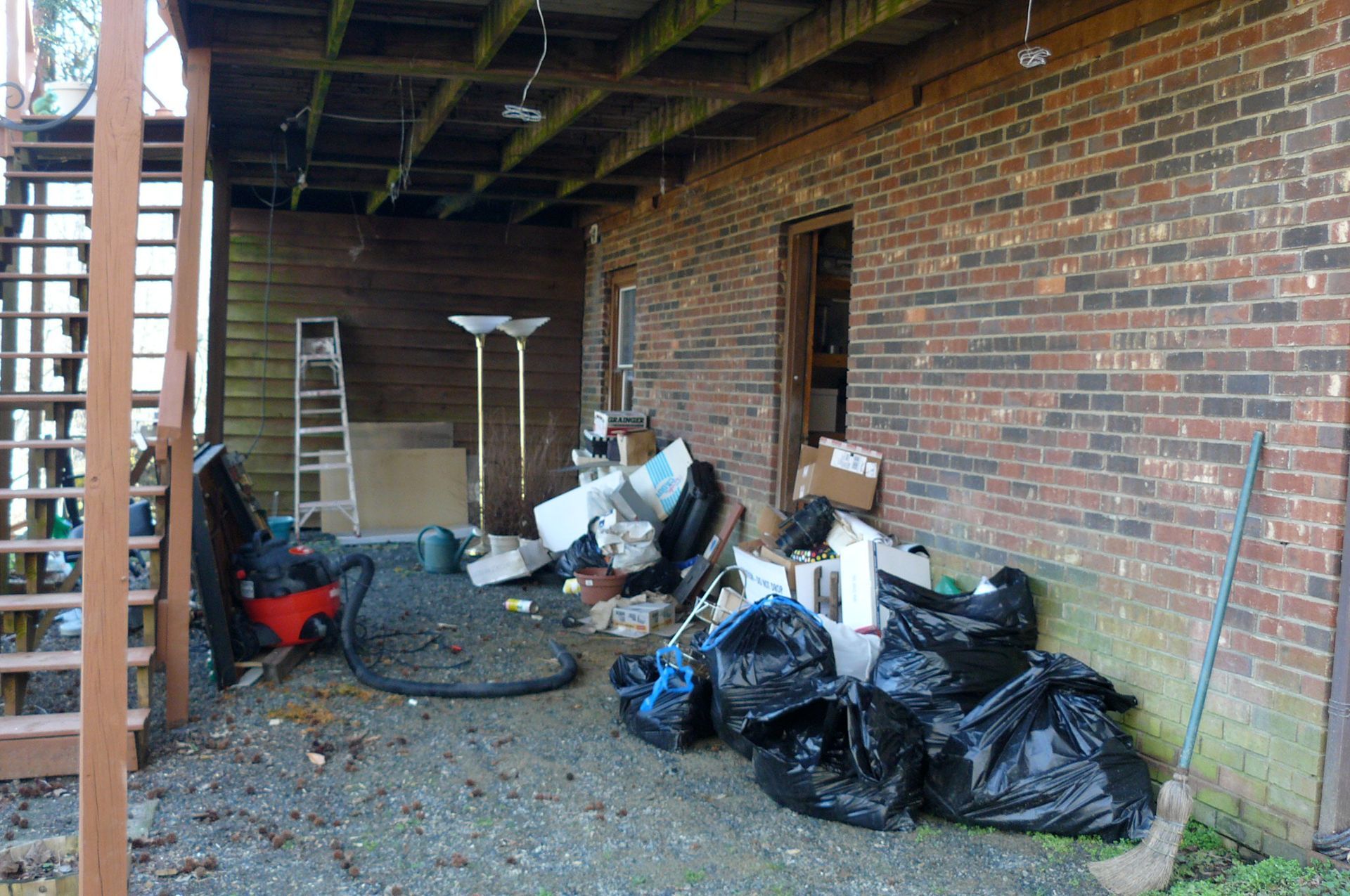 Exterior of a brick building with a wooden porch and stairs, cluttered with trash and debris.