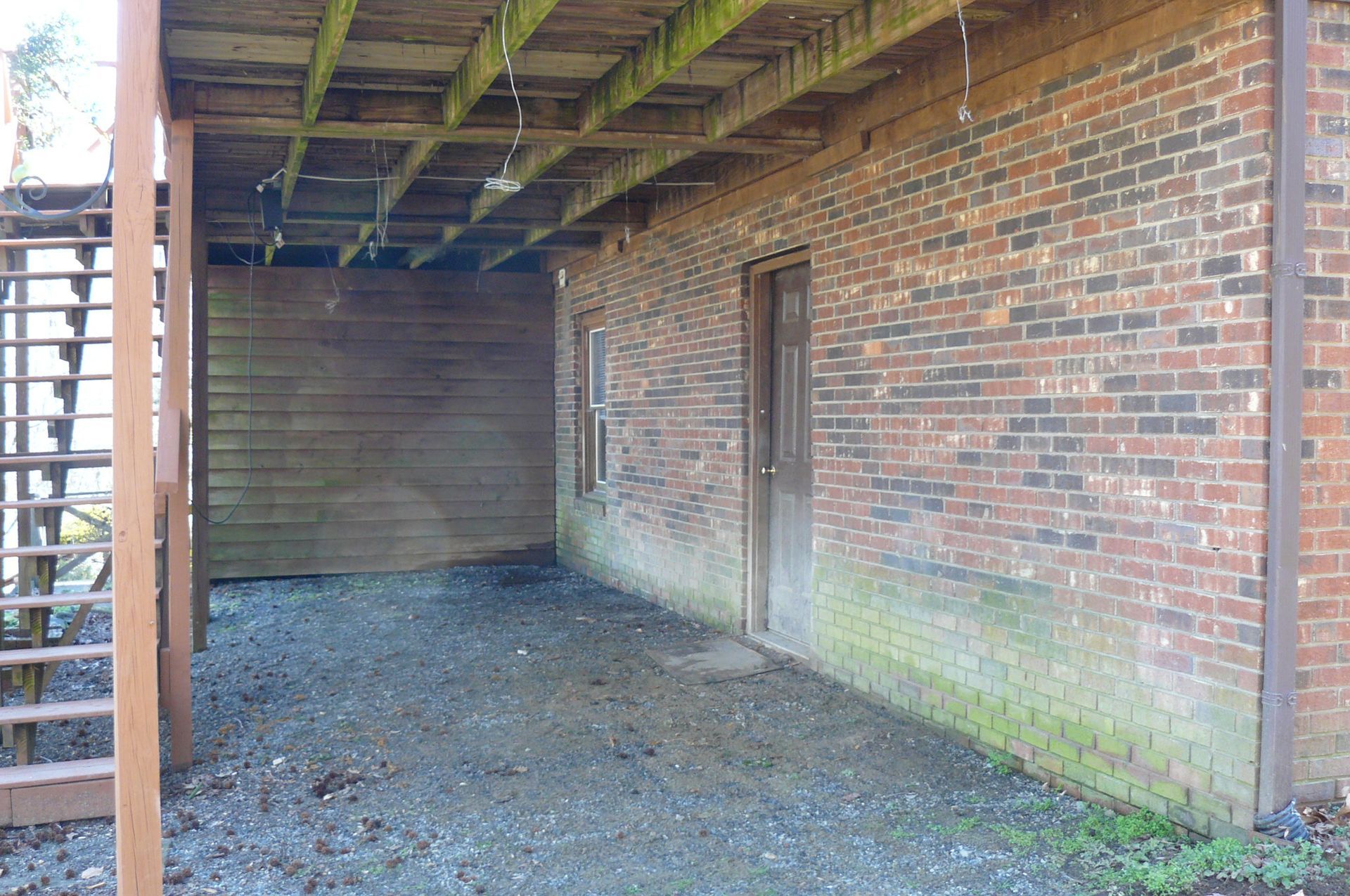 Underneath a wooden deck, a gravel patio sits next to a brick building with a wooden door and small window.