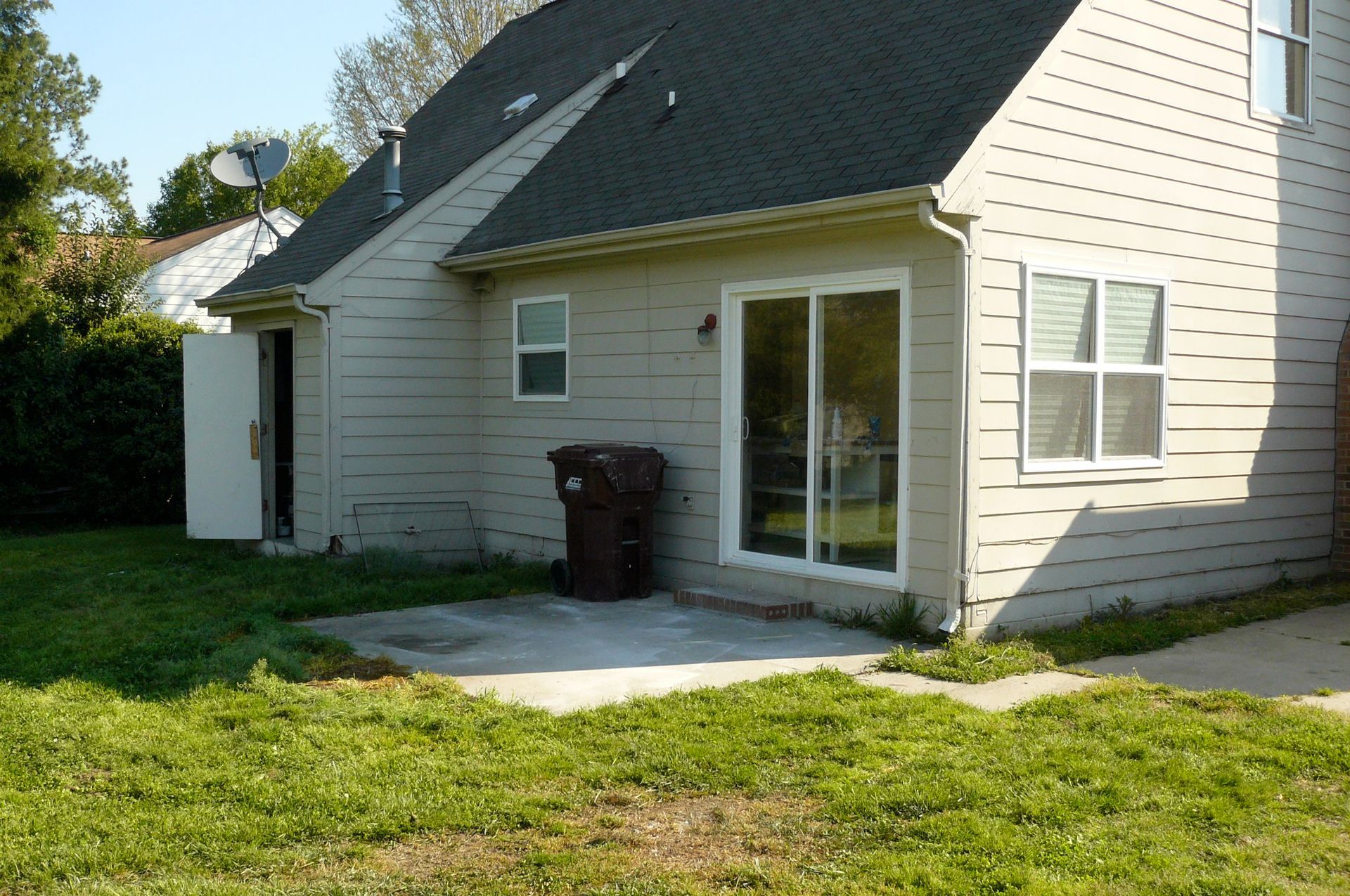 Backyard view of a beige house with a sliding glass door, patio, open shed door, and a brown trash can on the patio.