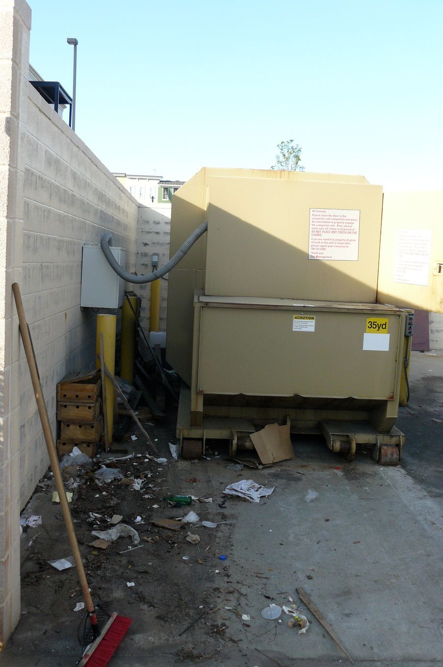 A large beige trash container is surrounded by garbage and a broom in an alleyway.