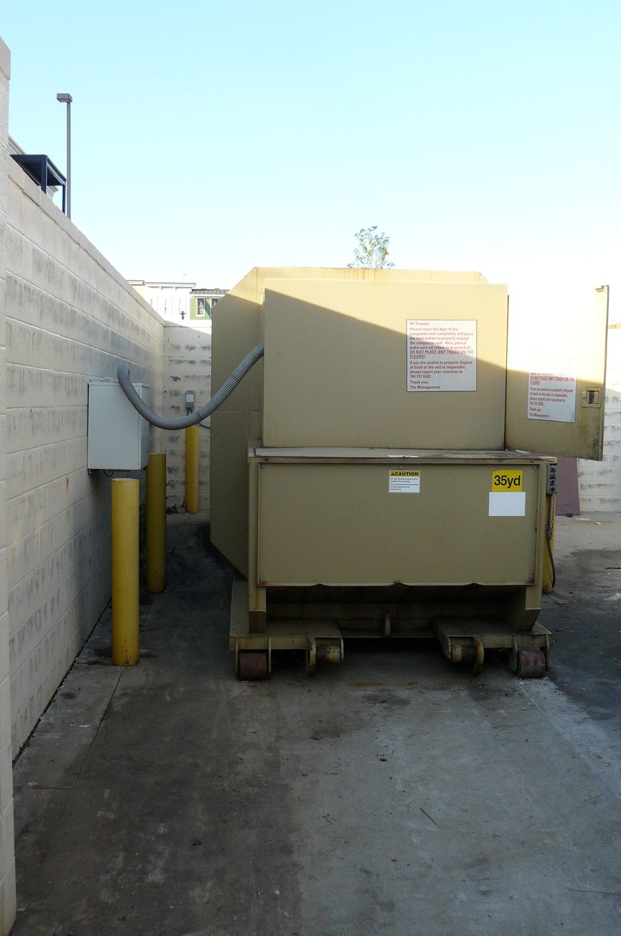 Large beige dumpster in an alley, between a concrete wall and yellow bollards.