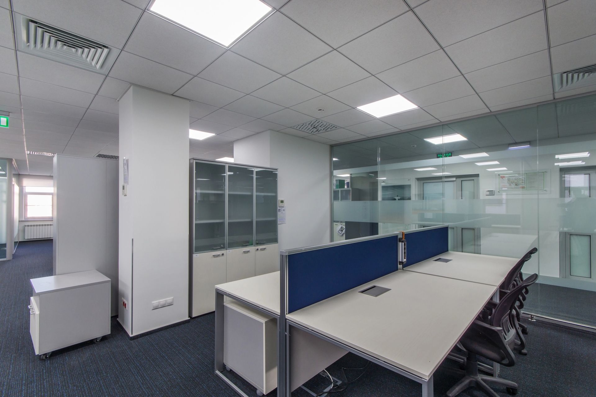 Office interior with desks, cabinets, and glass walls. Blue carpet, white walls, and bright ceiling lights.