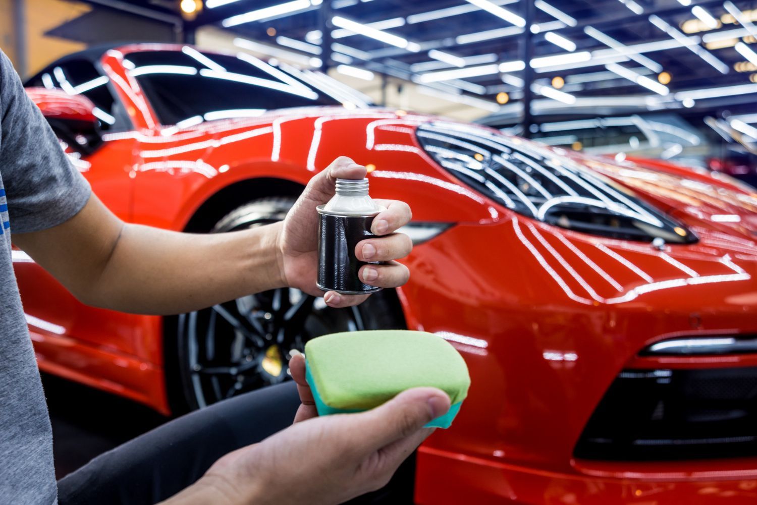 A man is holding a spray bottle and a sponge in front of a red car.