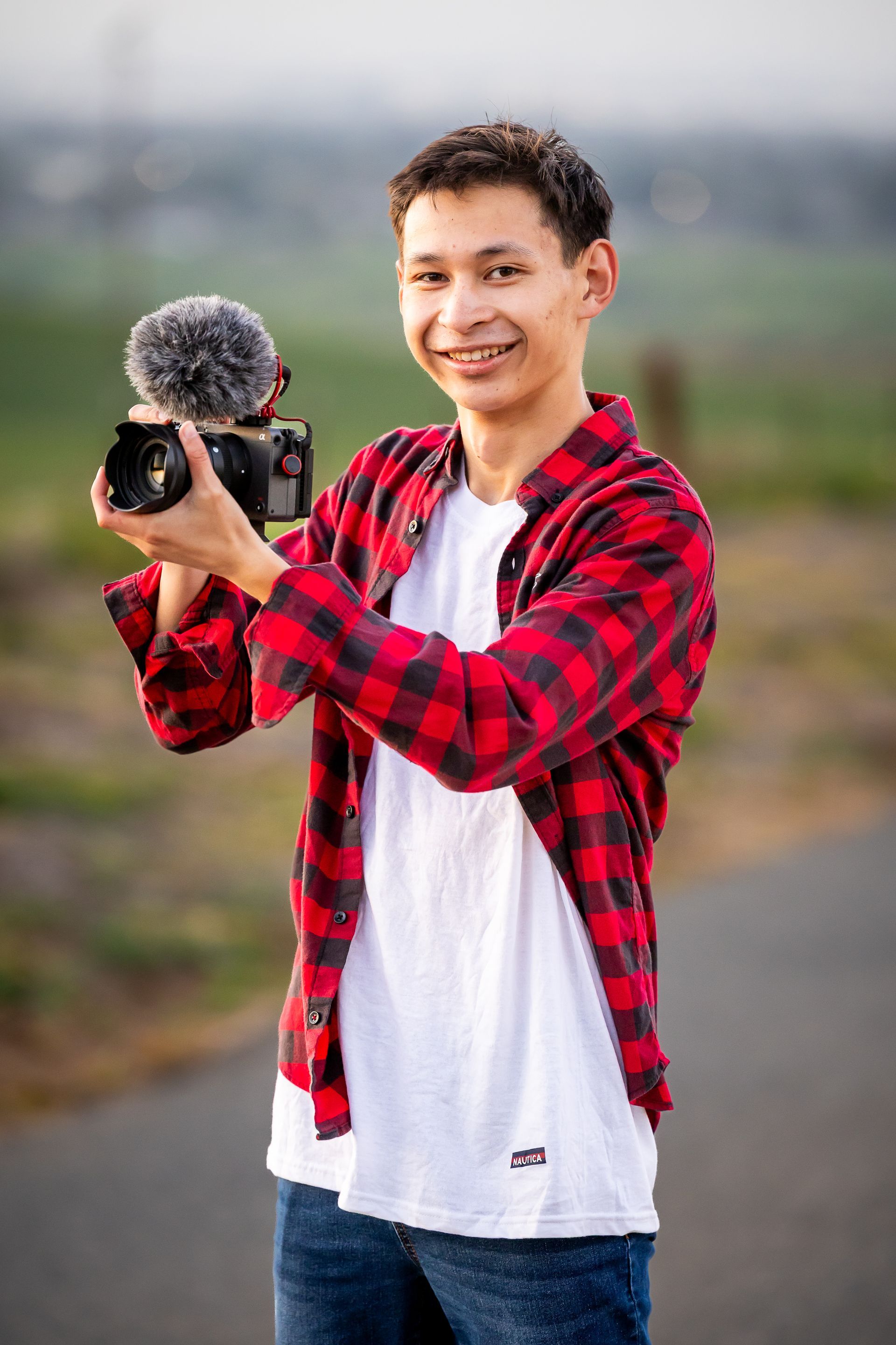 Man holding camera with microphone, wearing red plaid shirt. Outdoor setting.