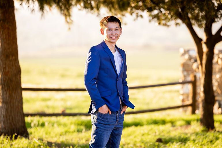 Man in blue blazer and jeans smiles outdoors, hands in pockets, near trees and fence.