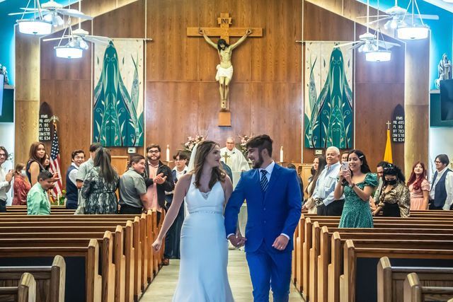 Newly married couple walking down church aisle, holding hands. Guests applaud. Crucifix on wall.