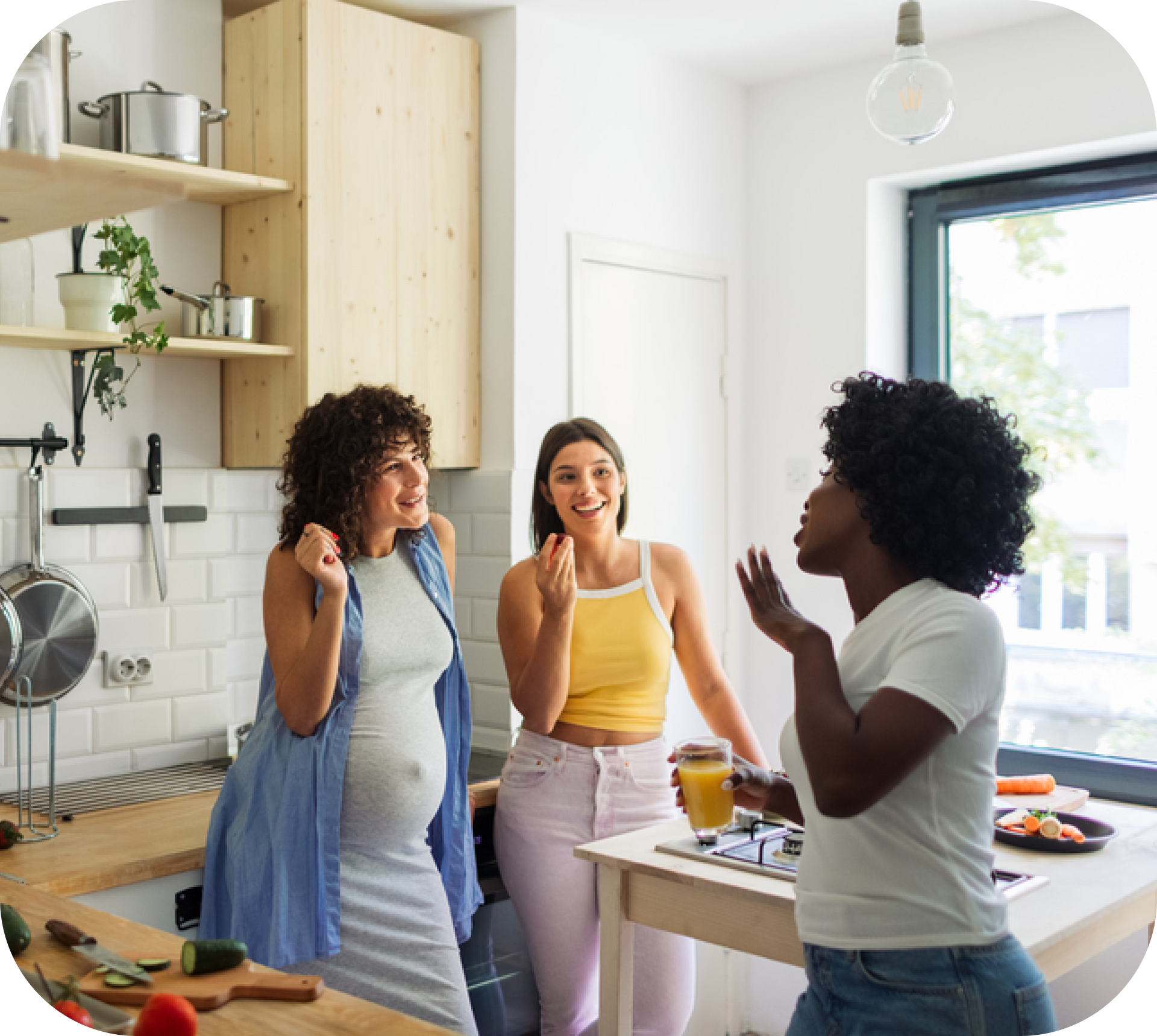 Three women are standing in a kitchen talking to each other.