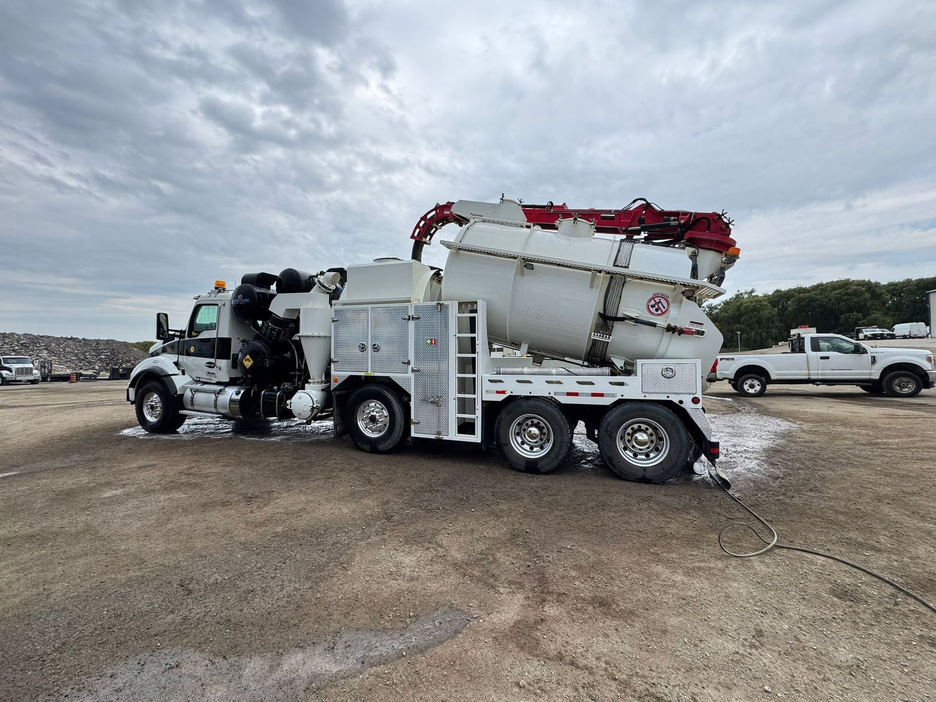 Concrete mixer truck parked on gravel, under cloudy sky. White truck with red details, with a white pickup truck in the background.