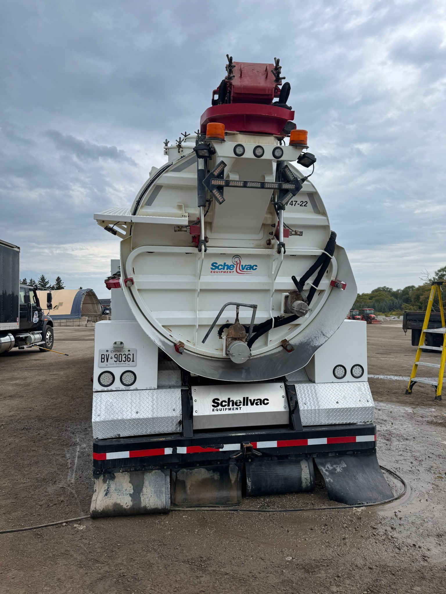 Rear view of a white vacuum truck with a red top structure, parked on a gravel lot under a cloudy sky.