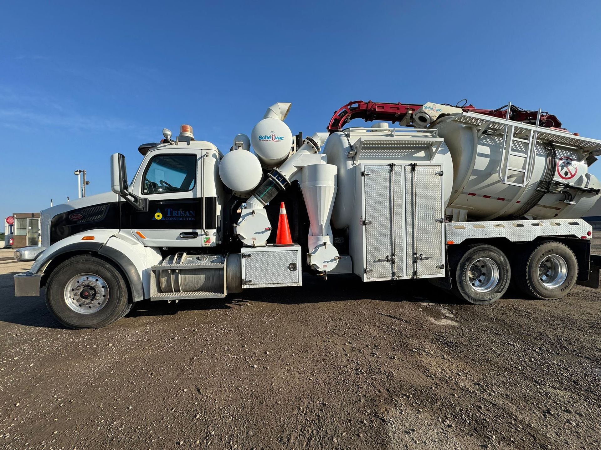 White vacuum truck parked on gravel, with large tank and suction apparatus. Sunny, clear sky.