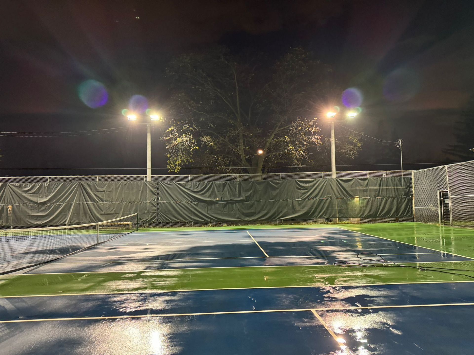 A wet, blue tennis court at night, illuminated by bright overhead lights. A tall fence surrounds the court.