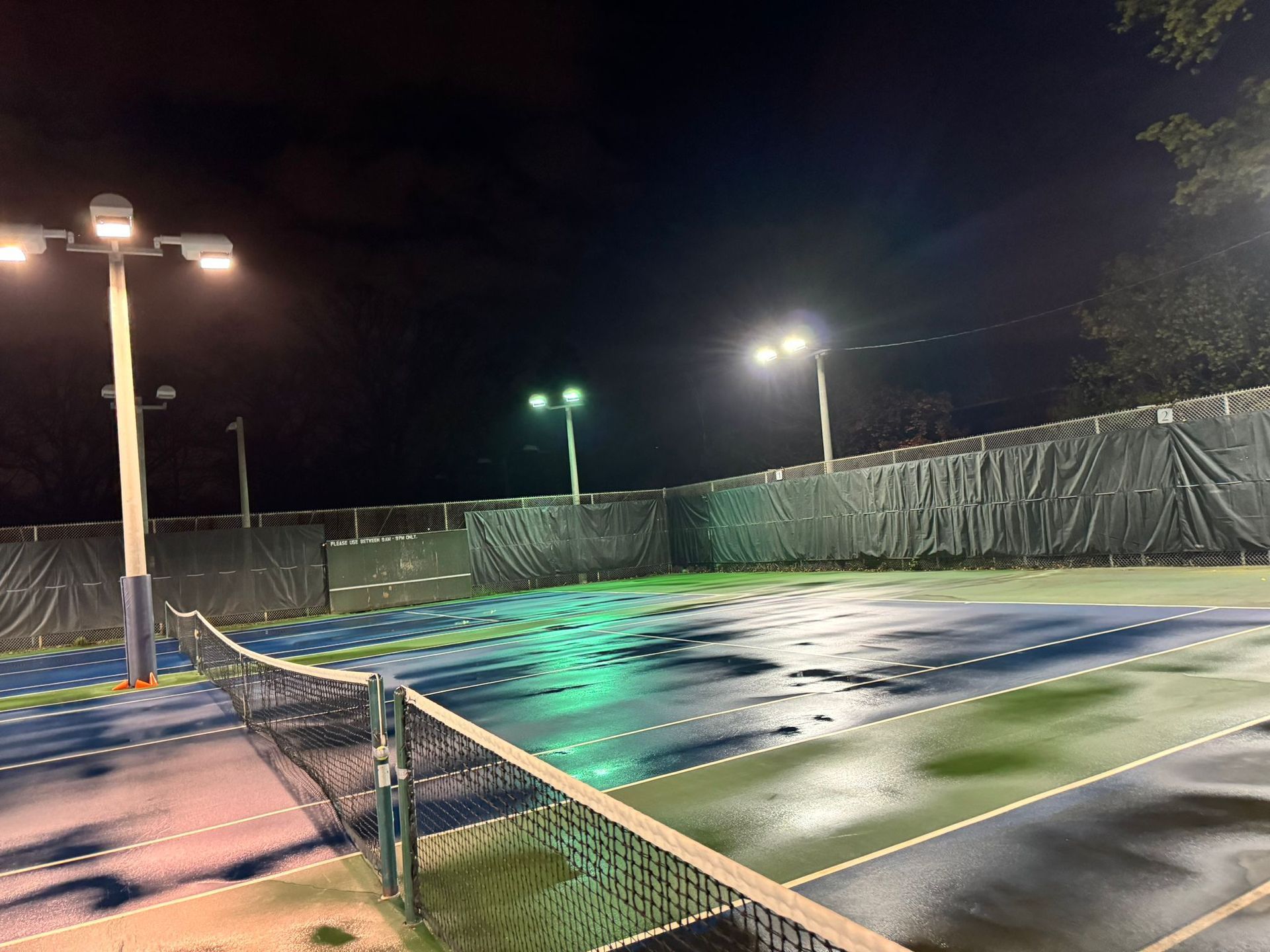 Nighttime view of a wet tennis court lit by overhead lights, surrounded by a black fence.