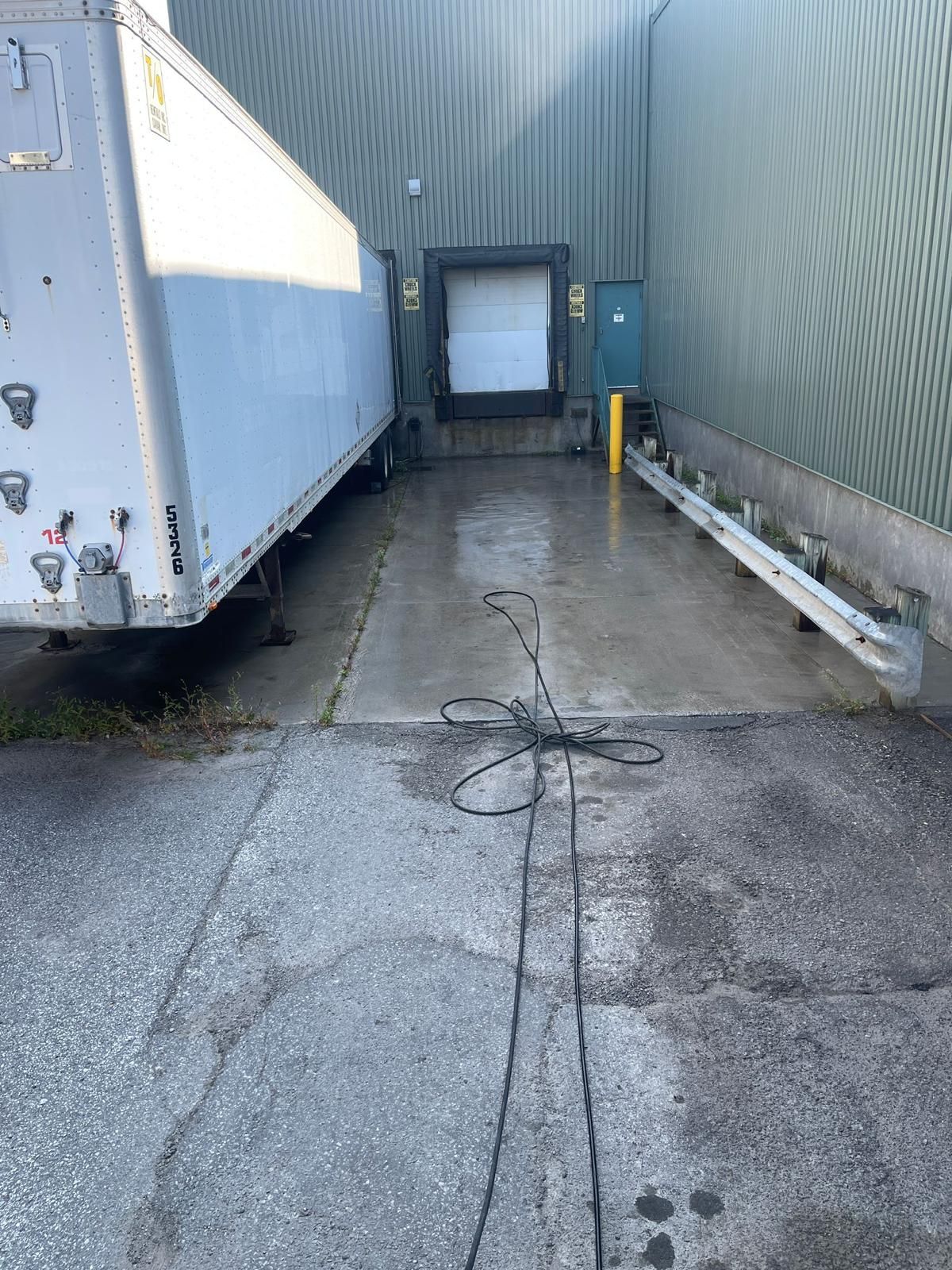 A semi-trailer truck backs into a loading dock. Black cable lies on the wet concrete.