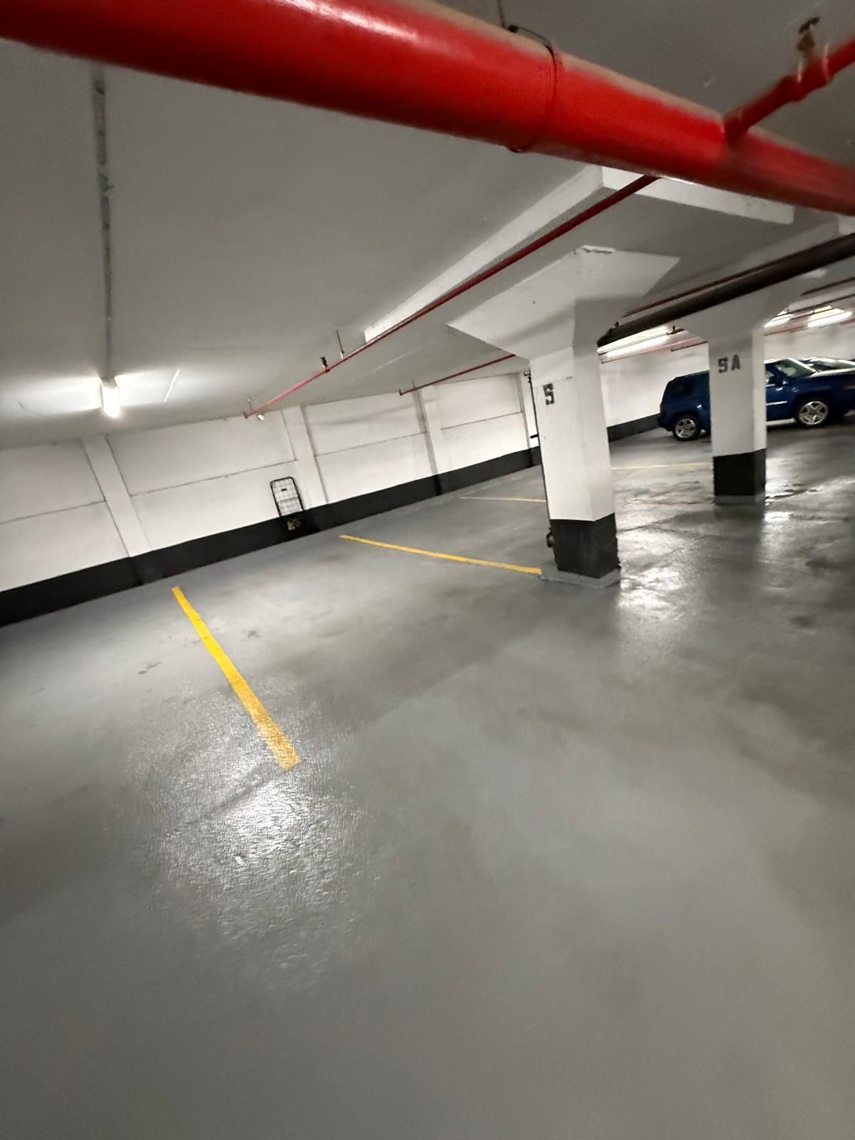 Underground parking garage with gray floor, white walls, and red pipes overhead. Yellow parking space lines and two cars visible.