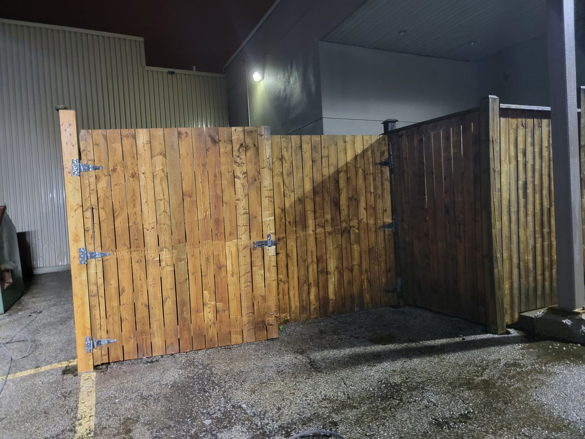 Wooden fence with gate open, against a wall. The wood is stained dark, set outside at night.