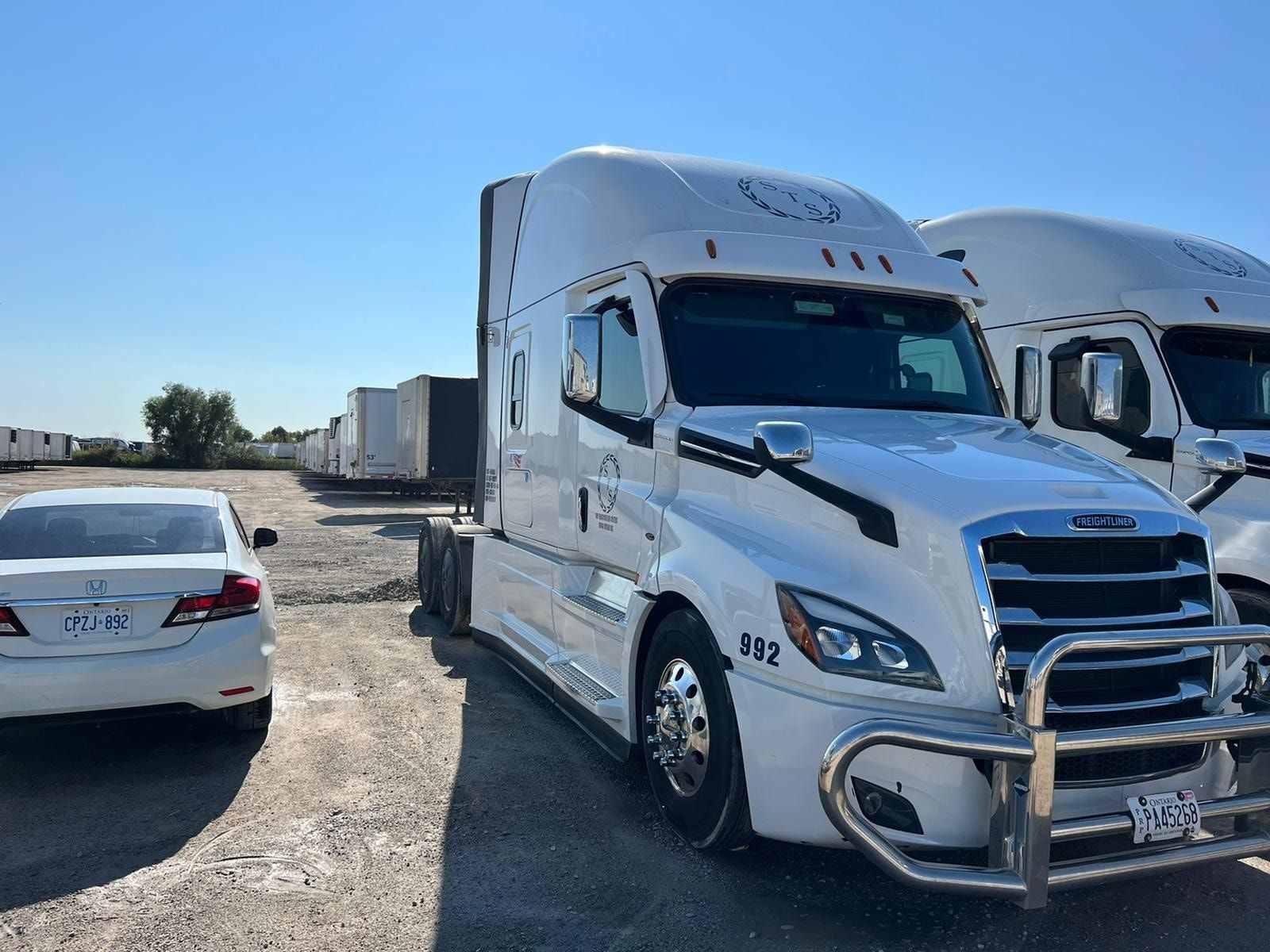 White semi-truck parked outdoors on a sunny day, with a car in the foreground and a row of trailers.