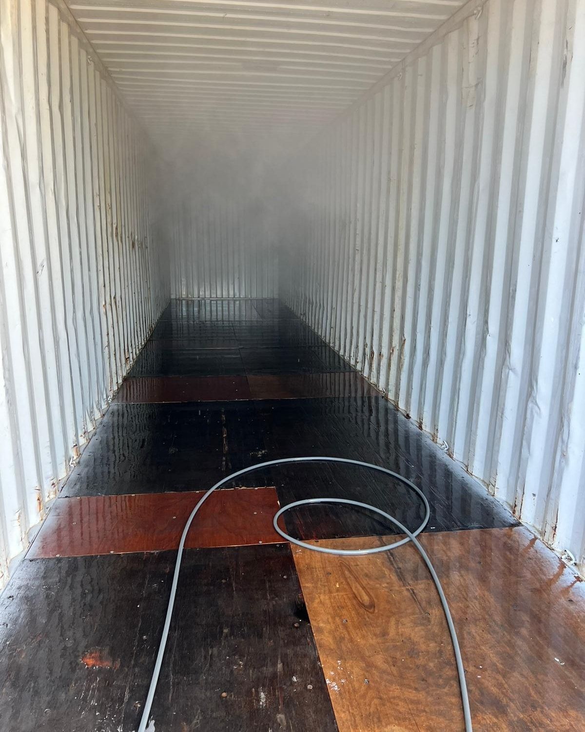 Inside a shipping container, the floor is wet with a hose coiled in the foreground; steam or mist obscures the far end.