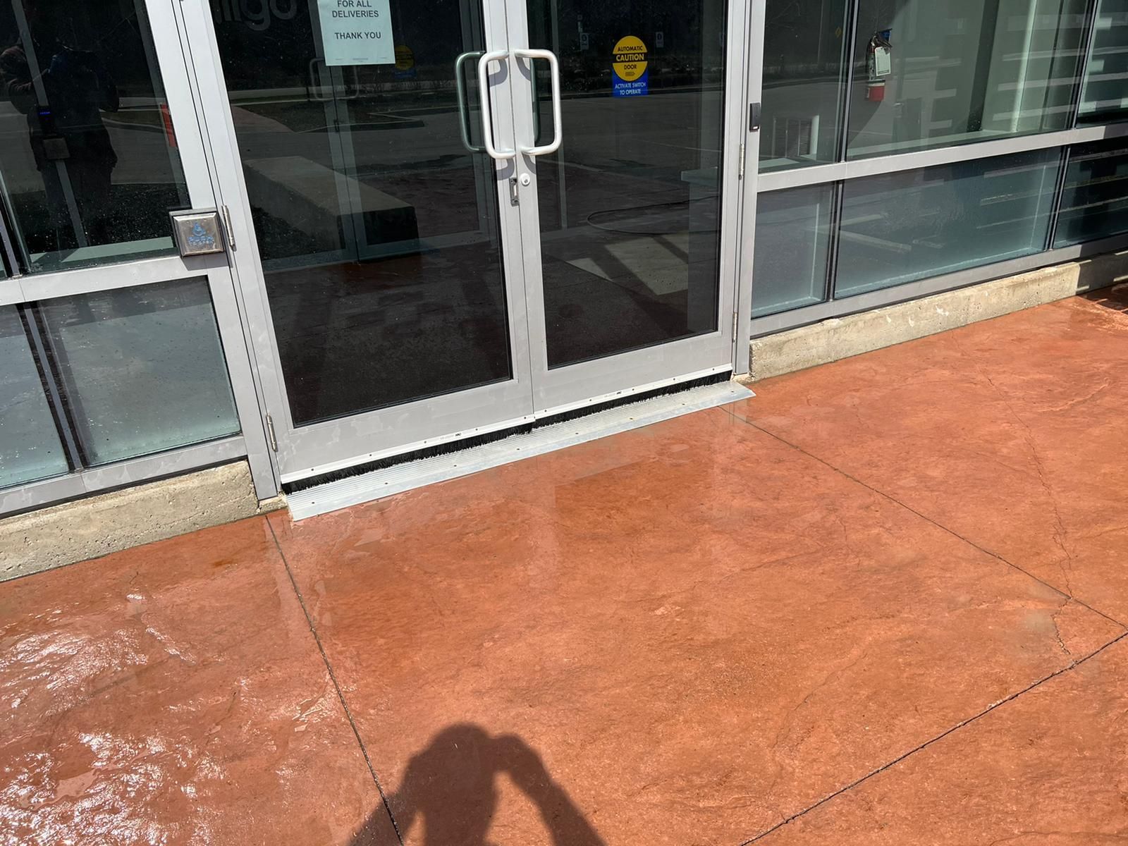 Exterior view of a building's entrance: glass doors, brown concrete surface, and bright sunlight.