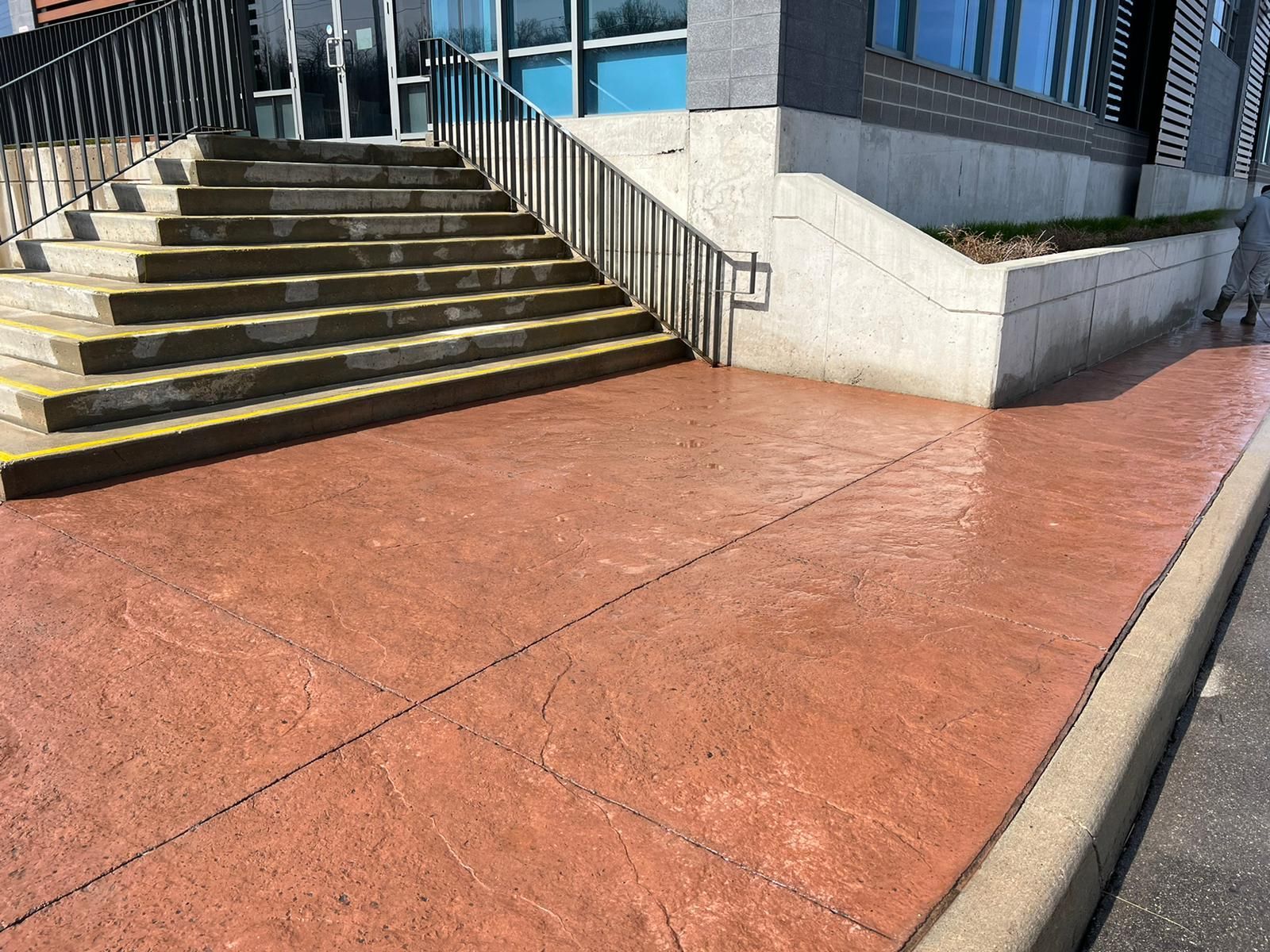 Red concrete walkway leads to stairs beside a building with a glass facade.