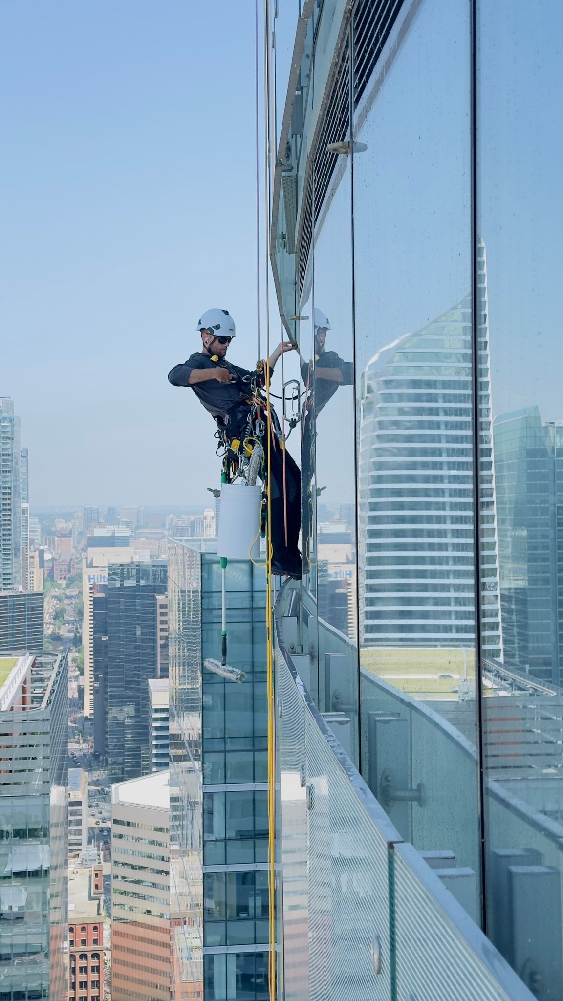 Window washer rappels down a skyscraper, cleaning glass. City skyline in background, blue sky.