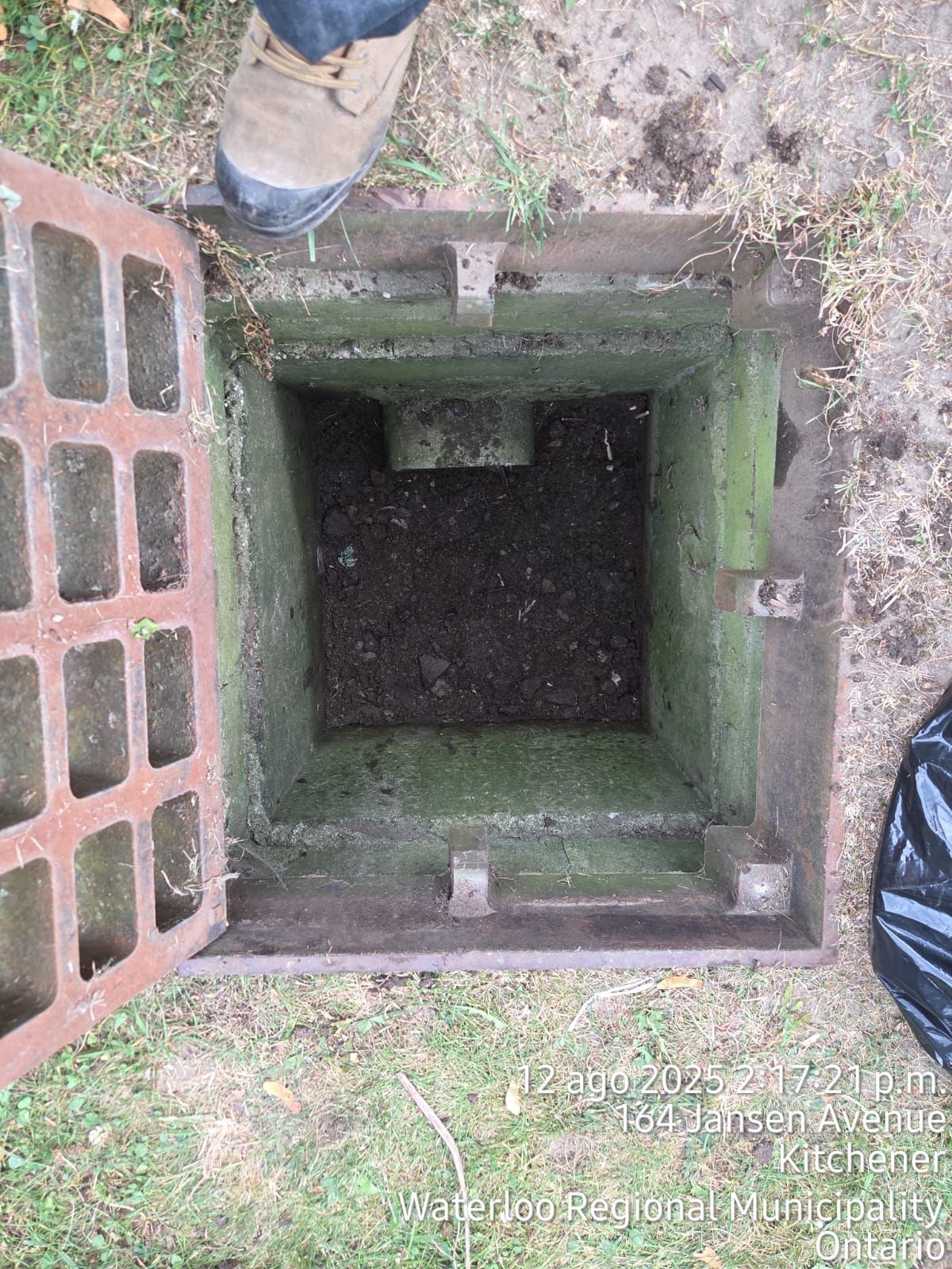 Open manhole with a square opening. A person's boot is visible. The manhole is on grass.