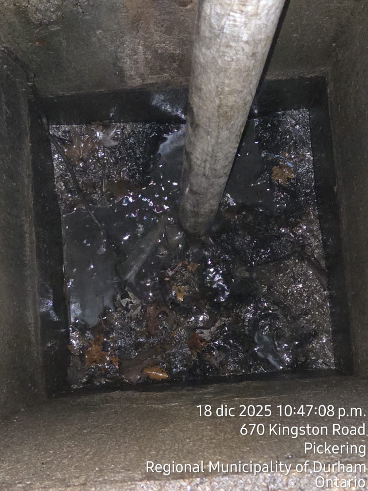 View down into a manhole, filled with debris and a pipe; text indicates location and date/time.