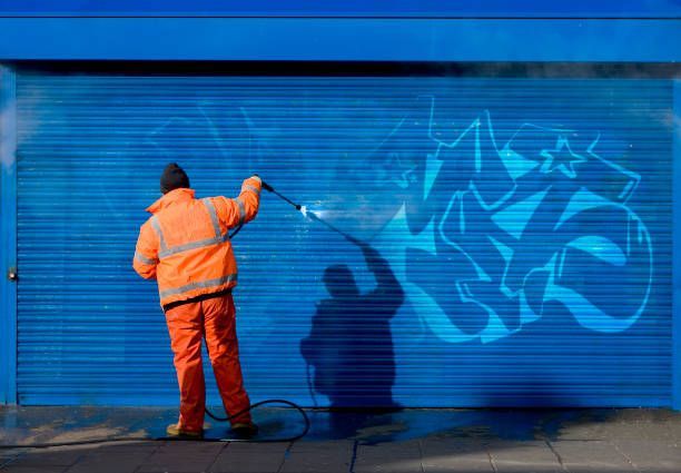 Person in orange cleaning blue graffiti from a blue garage door with a pressure washer.