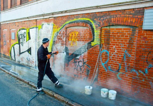 Man power washing graffiti from a red brick wall on a street. Three buckets sit nearby.