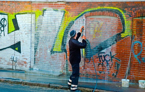 Person removing graffiti from a brick wall with a long-handled brush and water.