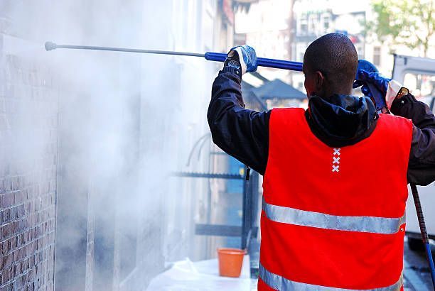 Person in orange vest pressure washing a brick wall. White mist fills the air.