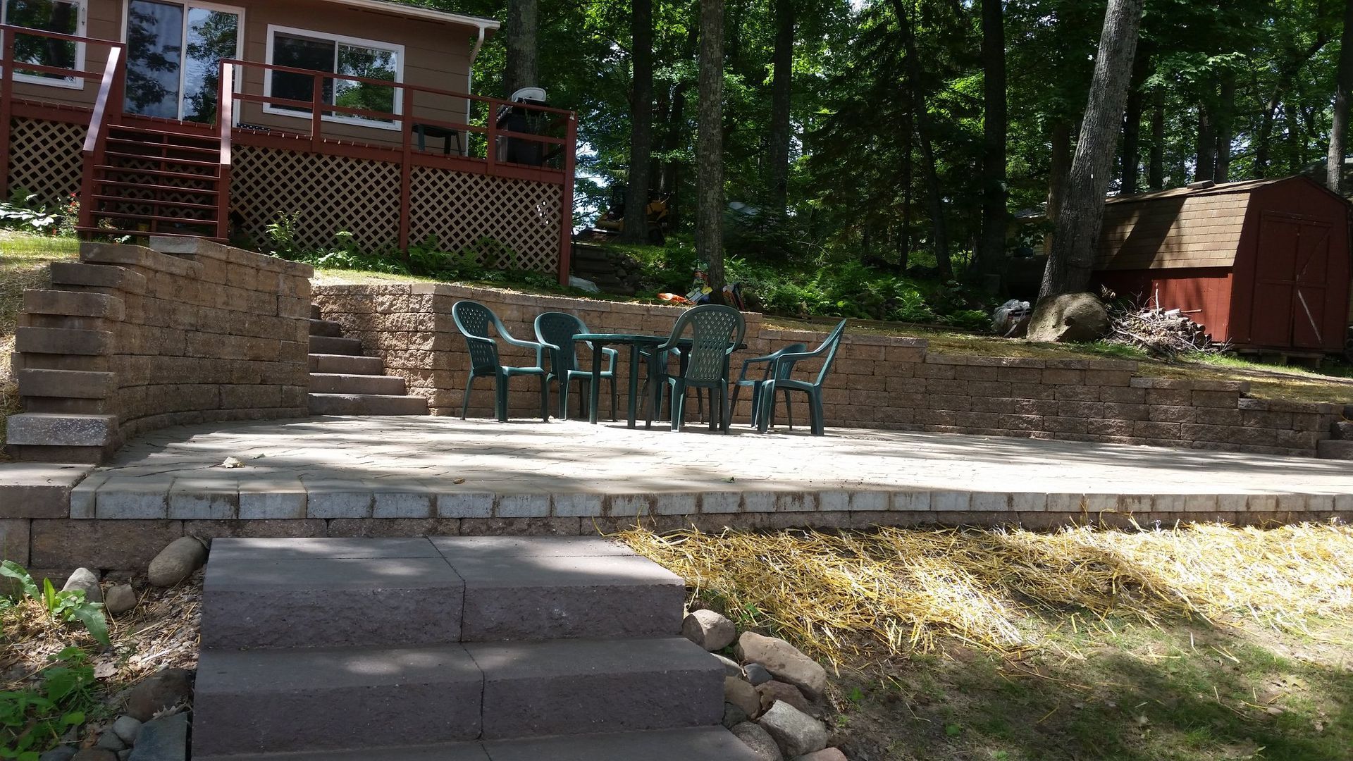 Stone patio with table and chairs, near a brown cabin and shed, in a wooded area.