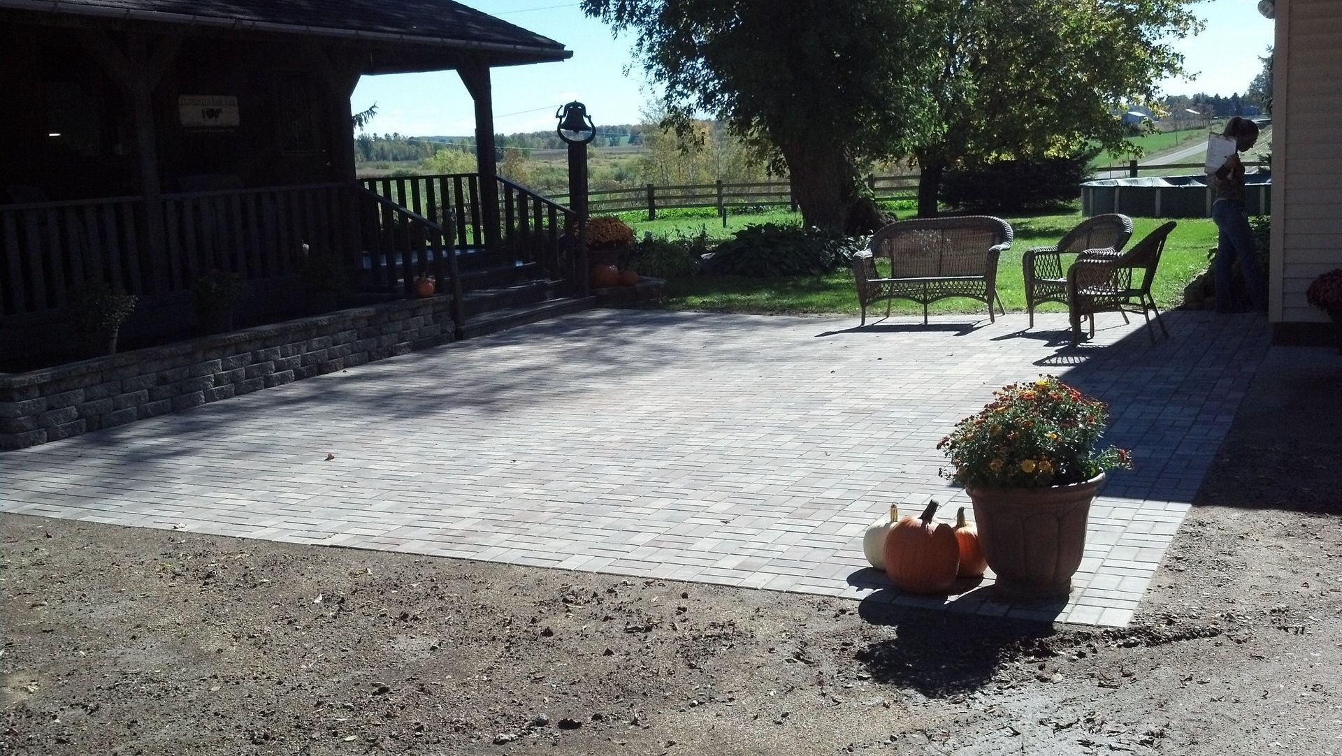 Brick patio with seating, potted flowers, and a pumpkin, near a building with a porch and a view of a grassy field.