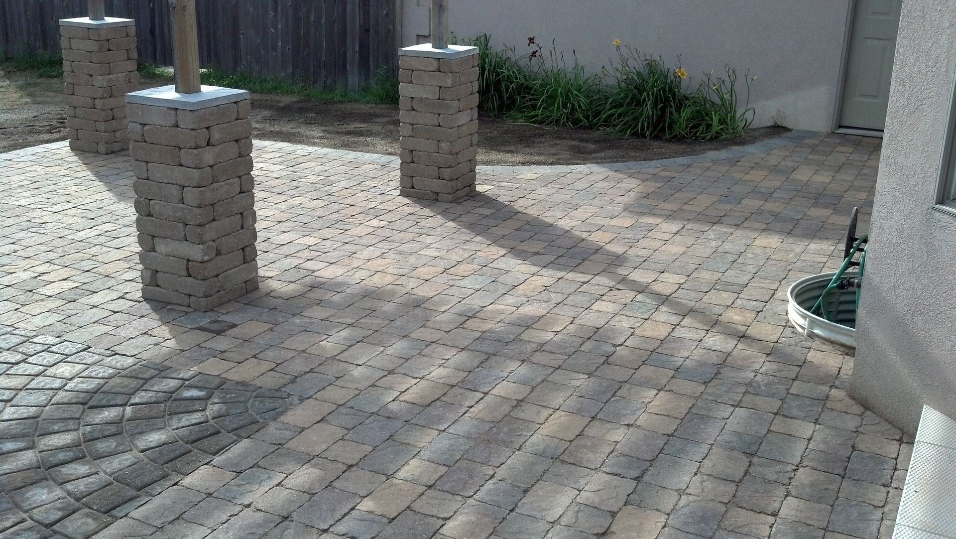 Stone patio with brick columns and a wooden support, next to a building and fence.