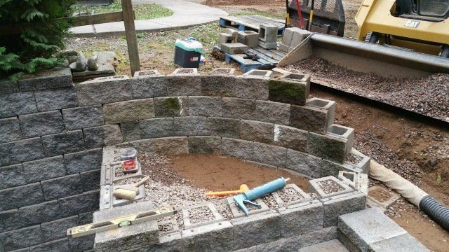 Stone retaining wall construction in progress. Blocks being laid in a curved design, sand visible.