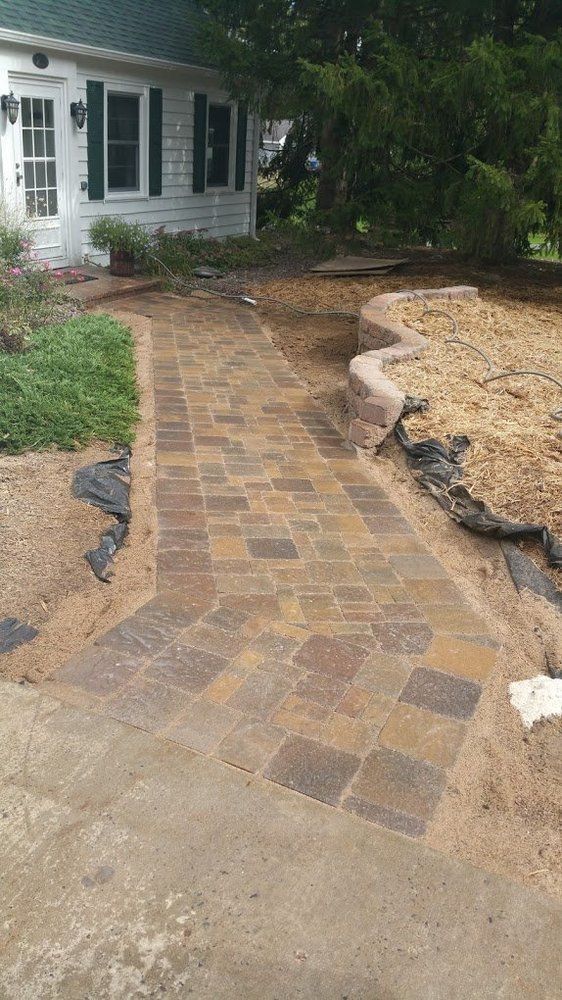 A brick walkway curves past a white house with green shutters, surrounded by sand.