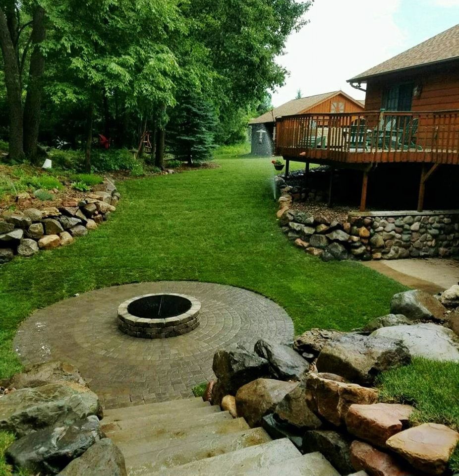 A backyard with a fire pit, rock walls, green grass, and a wooden deck.