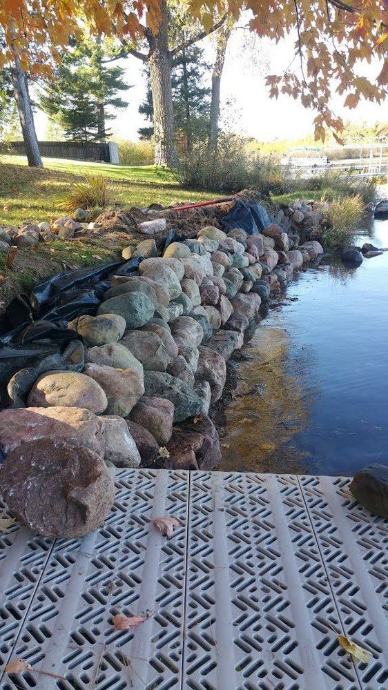 Rock retaining wall on a shoreline, beside a gray dock. Trees with fall leaves overhead.