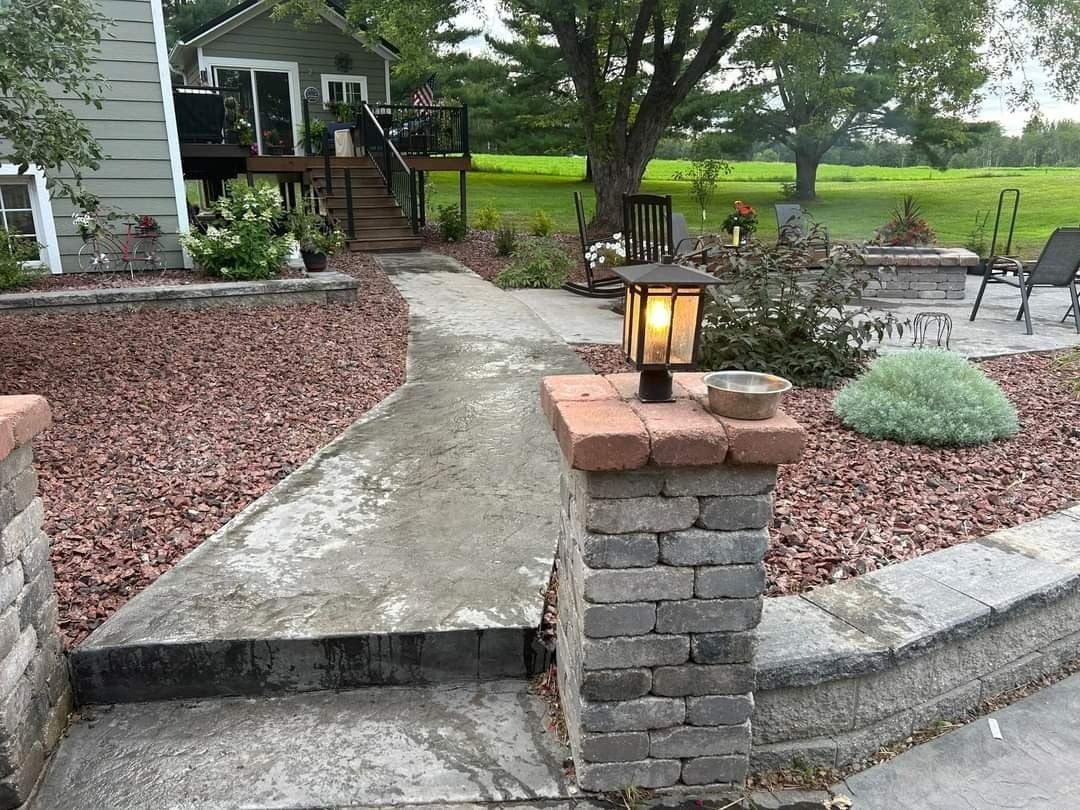 Brick pillars flank a concrete walkway with a solar lantern, leading to a house with a deck. Green lawn in the background.