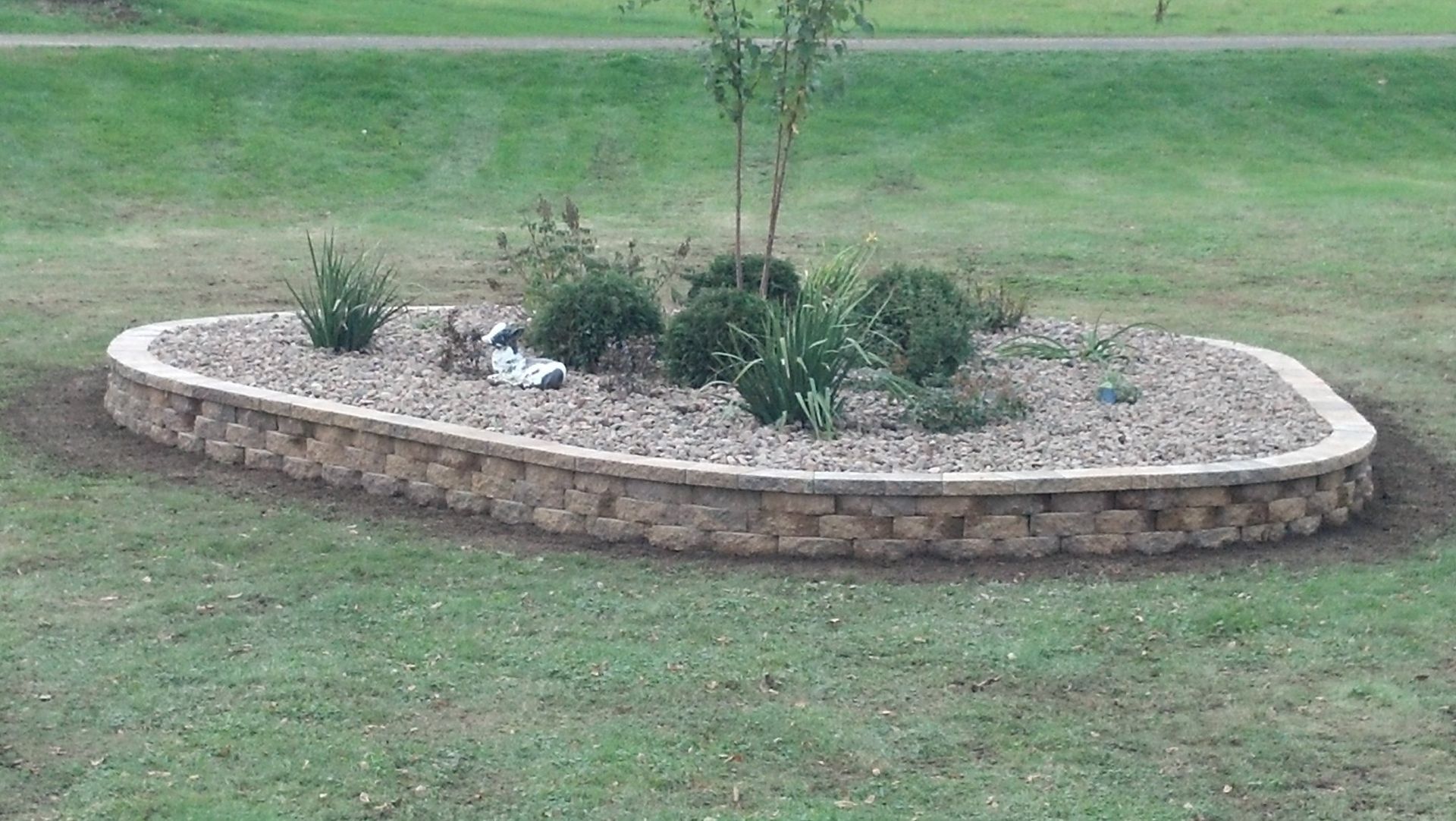 Raised stone flower bed with gravel and plants, in a grassy area.