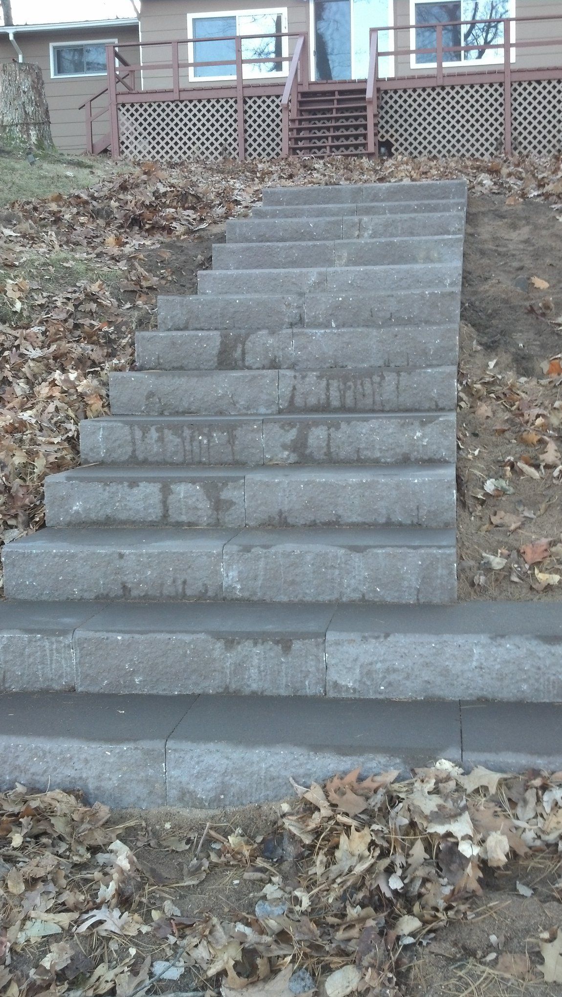 Concrete steps leading up a hillside to a house with a deck. The ground is covered with fallen leaves.