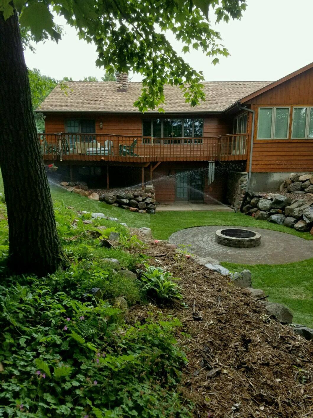 Wooden house with deck, green lawn, fire pit, and trees.