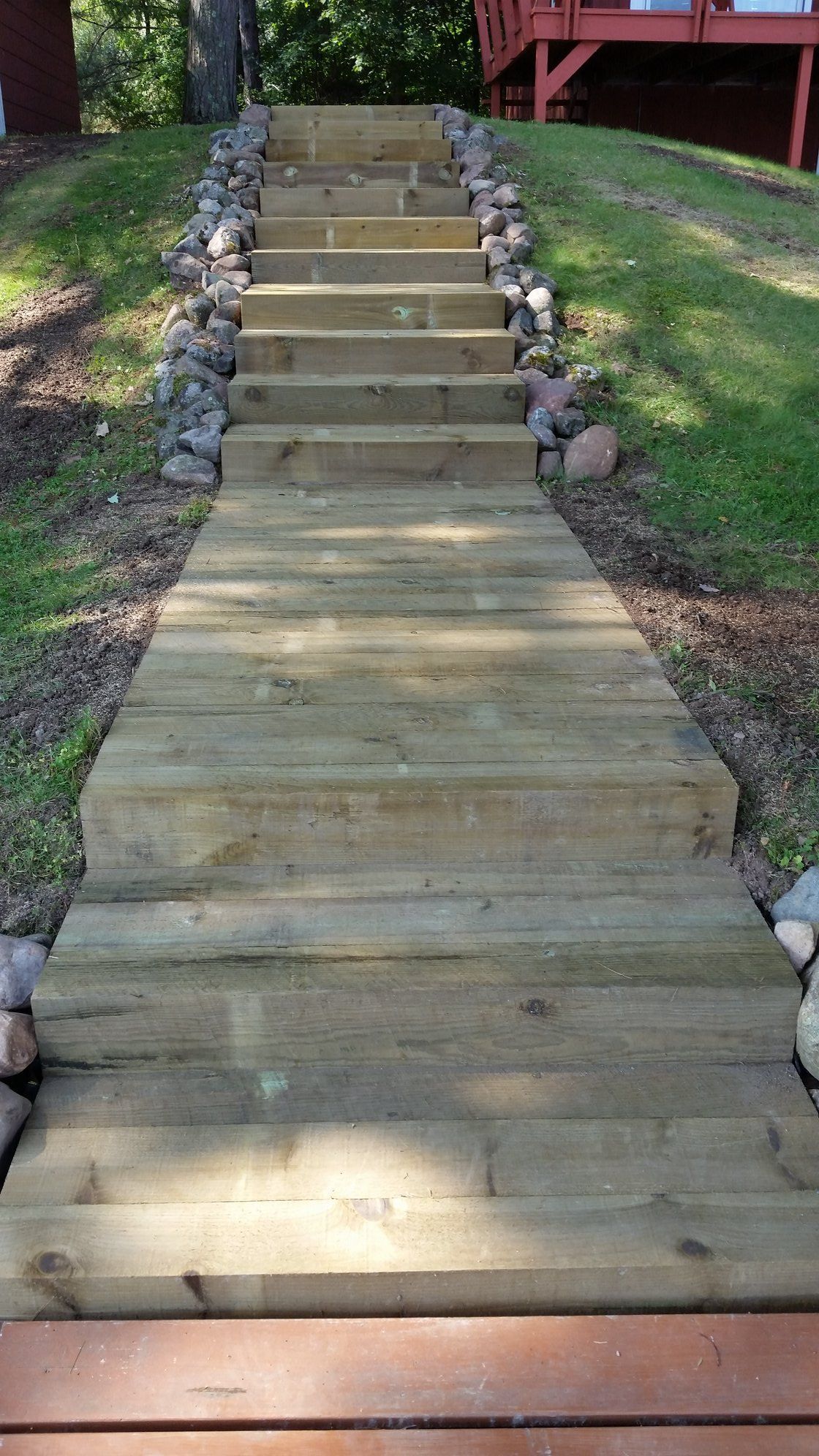 Wooden stairs ascending a grassy hill towards a house; large rocks border the stairs.