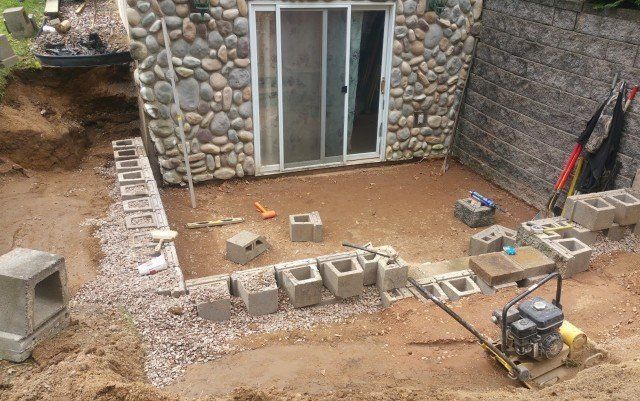 Construction of a patio next to a building with stone exterior. Cinder blocks, gravel, and a compactor are present.